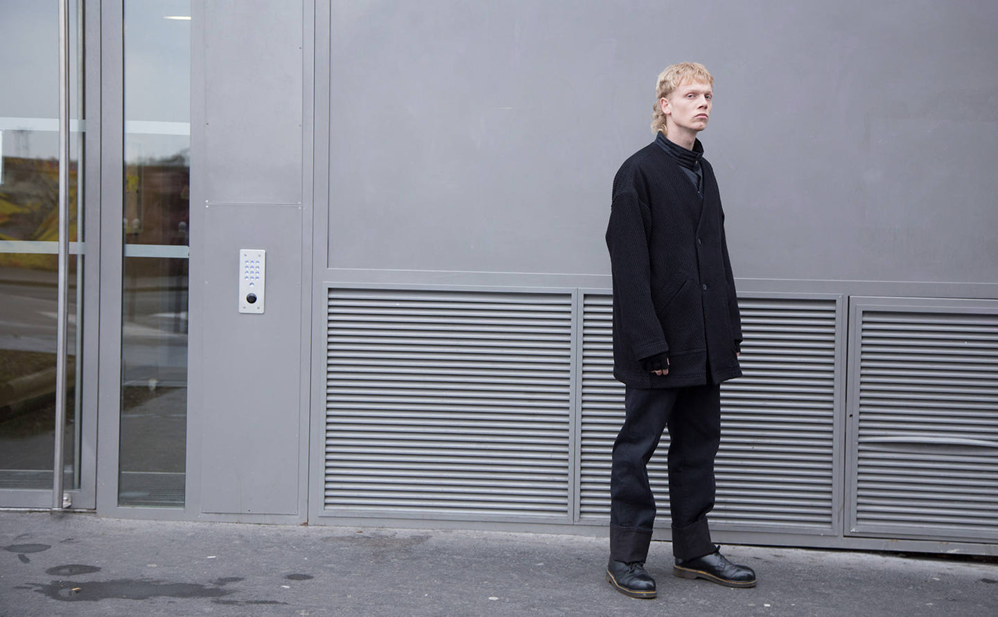 A model in an oversized black jacket and black denim standing before a grey shutter