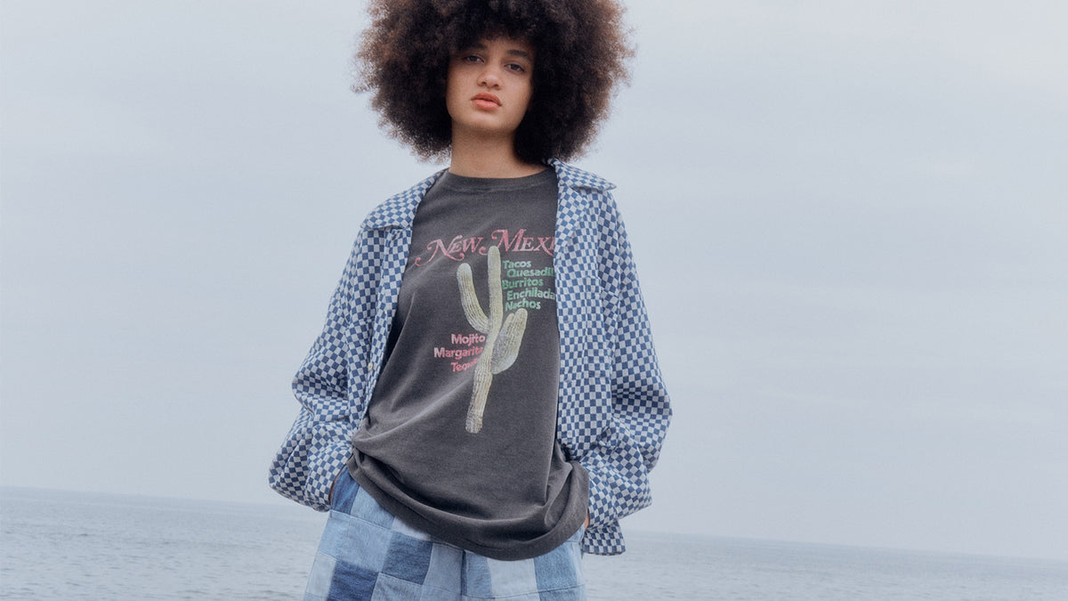 海を背に、プリントTシャツにギンガムチェックのシャツとパッチワークデニムを重ねたモデル