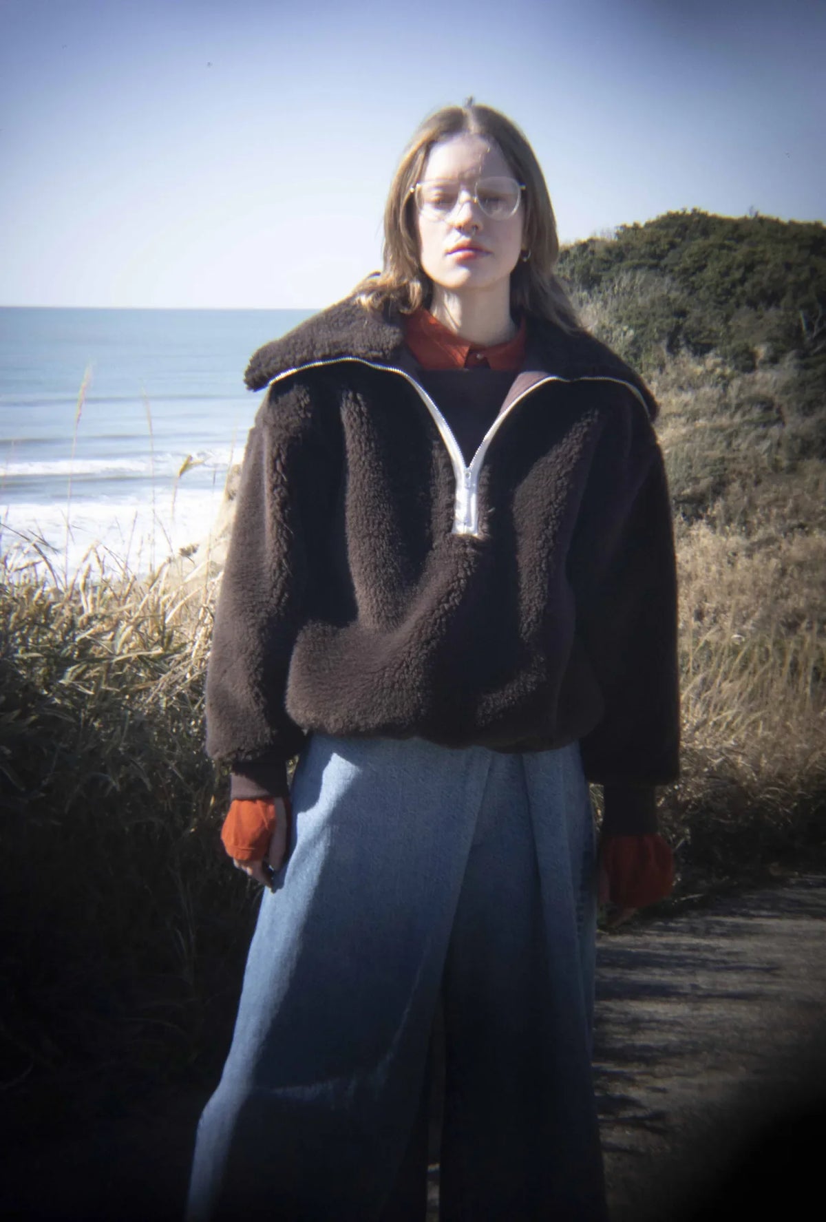 A model on a coastal path in a dark brown fleece half-zip and pale wide-leg denim, with the sea behind