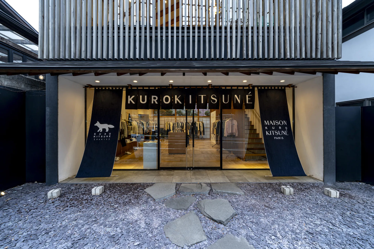 The Maison Kitsuné Daikanyama storefront at dusk, KUROKITSUNÉ lettering above the glass front and stepping stones across the gravel forecourt